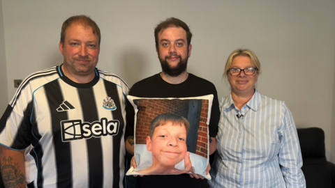 Craig Anderson, who is wearing a black and white Newcastle United football shirt, stands next to Aaron's brother, Lewis, who is wearing a plain black shirt and is holding up a cushion. The cushion has a picture of Aaron on it, which shows him smiling and giving a thumbs up. On the other side of Lewis is Aaron's mother Donna, who is wearing a blue and white striped T-shirt.