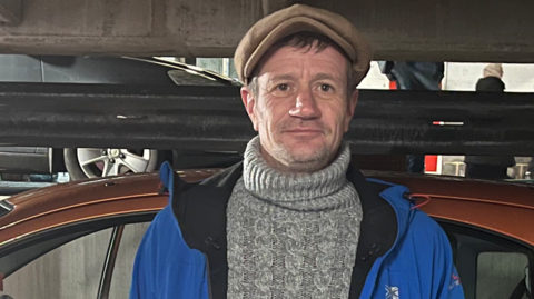 A man is standing next to an orange car. He is wearing a grey knitted jumper, a blue waterproof coat and a beige flat cap and is smiling at the camera. 