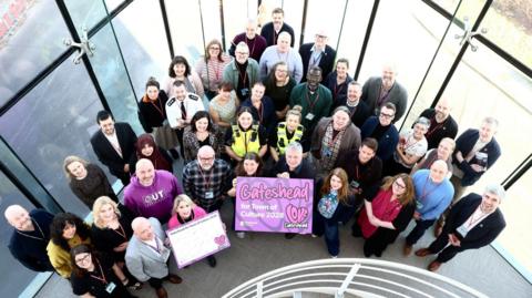 An aerial shot of a group of people standing in the corner of a building with windows behind them. Two people at the front have a purple poster saying Gateshead and a love heart.