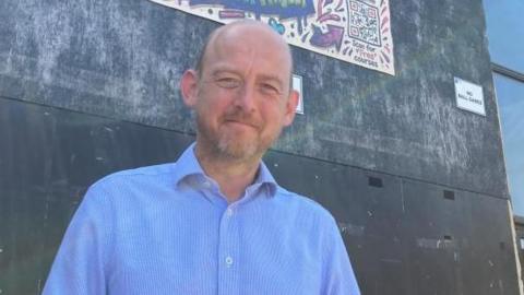 A man is standing in from a building. He is wearing a blue shirt and is smiling. Behind him is a brightly coloured sign which says "The Barn".