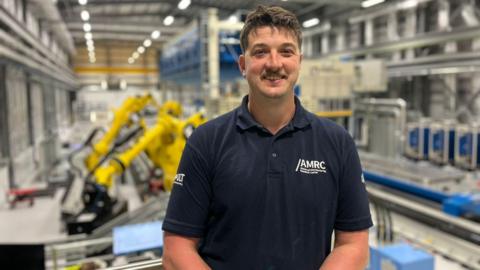 A man with short brown hair and a moustache is wearing a navy blue polo top with the words AMRC and smiling. He is standing in a huge factory with yellow machines in the background