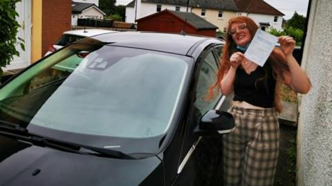 A woman with a driving test certificate next to a black car