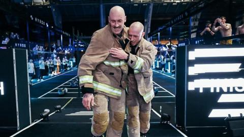Kieran and Andy across the finish line at Hyrox. Andy on the right of the picture leans into Kieran for support. Kieran is smiling in relief.