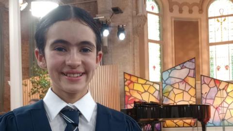 Zoe. She has dark hair tied back, wearing a navy school uniform, white shirt and navy striped tie. She is standing in a church, in the background there are stained glass windows, a black grand piano and studio lights.