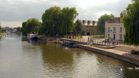 A picture of the river in Ely. A man is feeding swans in one section, a number of narrow boats are moored on the right hand side. Willow trees, a cream coloured building and a brown 3 storey Maltings building are in the background. 