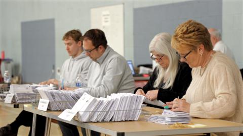 Two dark haired men in grey jumpers and two women, one with white hair, a black top and glasses and one with blonde short hair and a cream jumper counting ballot papers at a fold-down table, with three baskets of ballots in front of them