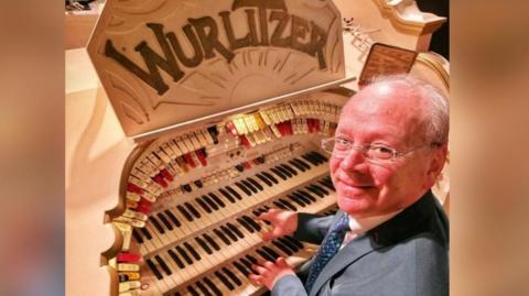 Phil Kelsall sits at the console of the Wurlitzer organ in Blackpool Tower Ballroom with his hands on the keyboards. He's wearing a dark grey suit, white shirt and blue tie.
