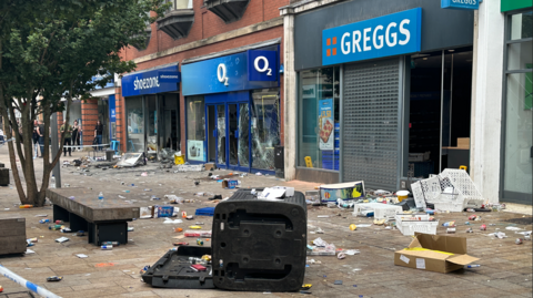 Damaged shops on Jameson Street in Hull. The stores are surrounded by police tape and rubbish and debris can be seen strewn across the pavement. Smashed windows can be seen at Greggs, O2 and Shoezone.