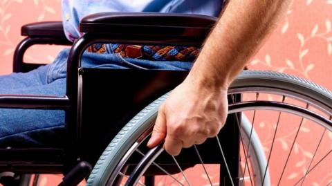 Close-up of an anonymous man sitting in a wheelchair. He is gripping one of the wheels with his left hand. He is wearing belted blue jeans and a light blue shirt.