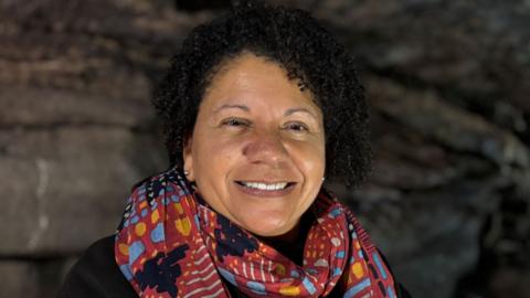 A middle aged woman with light brown skin and short curly dark hair. She is wearing a colourful scarf over a black long-sleeved top. She is smiling at the camera