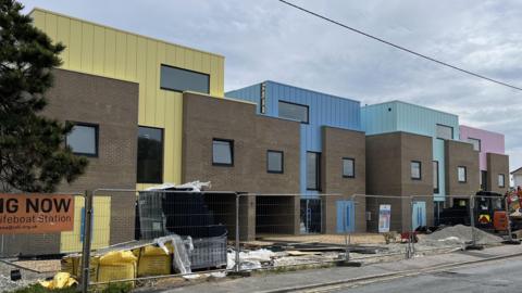 Four square houses behind construction fences. Each house is heavily accented with a different colour - yellow, blue, light green and pink.
