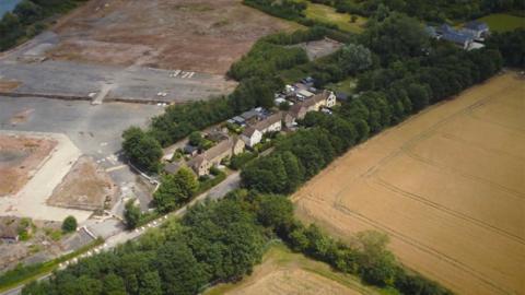 An aerial view of a row of 12 houses situated between fields and a former brickworks. The former brickworks is mostly made of concrete and there are arable fields to the front of the properties.