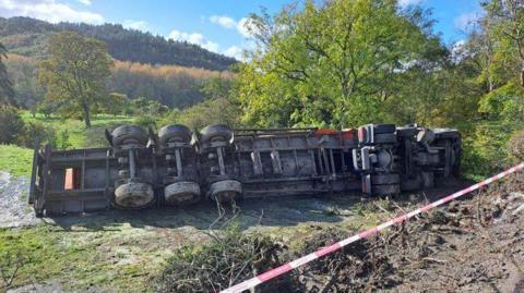 A large lorry is overturned on its side in a field. There is mud and water on the ground underneath it. The photo is taken from the side of a road, where you can see mud that has been disturbed, and red and white tape running across