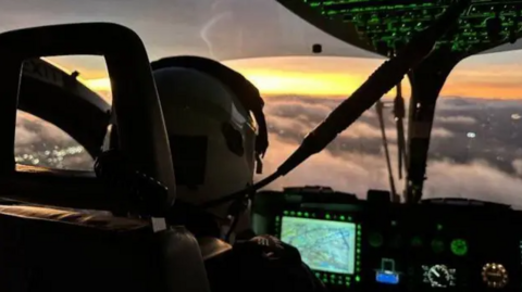 A close up of the inside of a police helicopter. The pilot is wearing a helmet and is sitting in front of a dashboard. The helicopter is high in the air above clouds.
