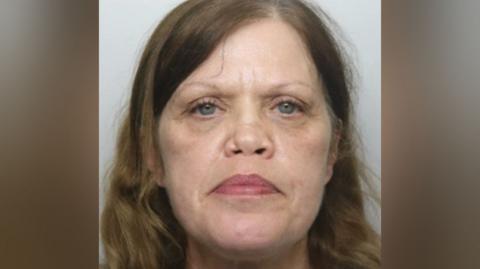 A mugshot of a lady with blue eyes and wavy mousy brown hair that is past her shoulders. She is looking straight into the camera.