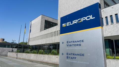 The exterior of the Europol building in the Hague - a dark blue sign with the Europol name in white text stands below a tall concrete building