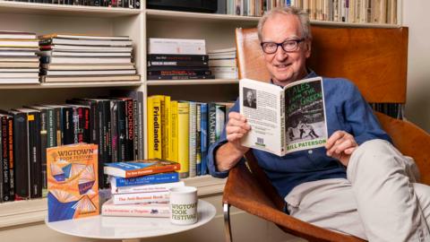 Actor Bill Paterson with grey hair and glasses in a blue shirt and light trousers reads a copy of Tales from the Back Green while sitting in a large, comfortable brown chair. There are books on the shelves behind him and a mug and books on a table beside him.
