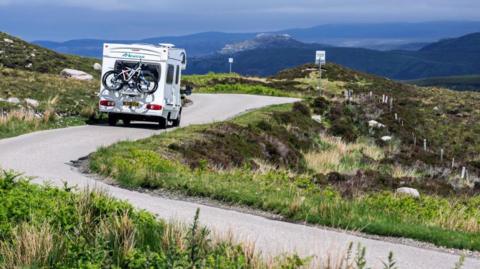 A white campervan with bikes attached to the back negotiates a winding Highland road. There are hills in the distance.