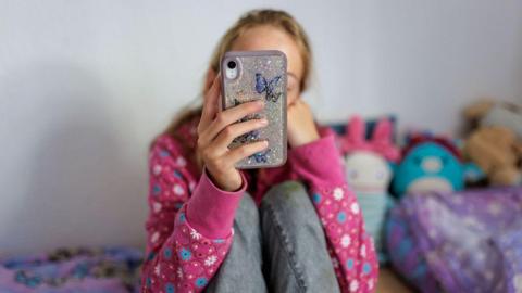 A anonymous young girl looking at her phone on her bed in a room with brightly coloured cuddly toys. She is wearing a pink flowery jumper and jeans, we cannot see her face.