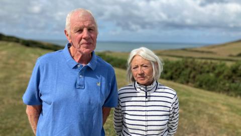 A man and a woman by the coast. There are rolling fields in the background and in the distance the sea and the North Devon coastline. 