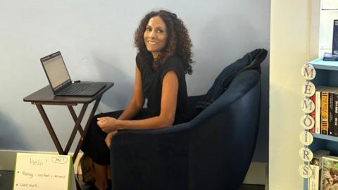 Kathryn Croft, a woman with long dark curly hair, wears a black sleeveless top and sits at a laptop on a desk in the Paper Moon bookshop in Guildford, smiling into the camera.