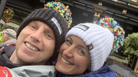 Andrew and Zoë pose for a selfie. They both have huge smiles on their faces and are dressed in coats and woolly hats.