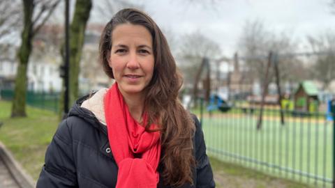 Emily Jennings. She is wearing a red scarf and a dark jacket. She has long brown hair. She is standing in front of a blurred background of a playground.