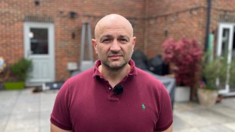 Man in red polo shirt and neutral expression stood on his garden patio with house in the background