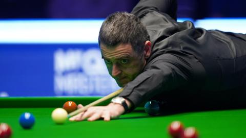 Ronnie O'Sullivan leaning on the snooker table about to hit the white ball. He is looking down at his cue and concentrating. He has short black hair and black clothes.