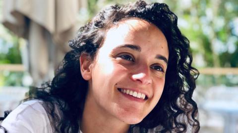 Emily Damari, a young woman with curly dark brown hair wearing a white t-shirt, smiles.