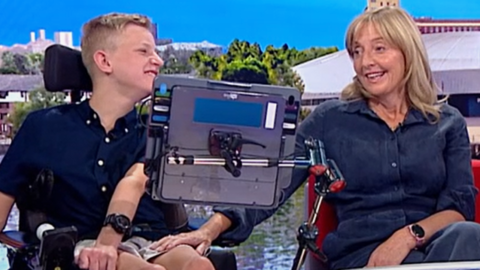 A young man in a wheelchair and with a tablet in front of him is next to a woman in a denim dress on a red sofa in a studio. She is holding on to his knee.