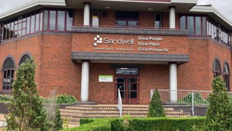 The entrance to Sandwell Council's headquarters - a brick building with a balcony over the entrance that says "Sandwell" in big white letters and "great people, great place, great prospects" next to it. There are steps leading up to the entrance and hedges and greenery in the foreground.