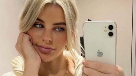 The photo shows Paris Wilson taking a mirror selfie indoors. They are holding a white iPhone with a dual‑camera setup in their right hand, and they are wearing a chunky striped knit top with a zip collar. Their hair appears to be light blonde and tied back loosely. A gold chain necklace is visible around their neck, and they are wearing a gold ring on their right hand. The setting looks like a simple, neutral-toned room, possibly a bedroom or changing area.