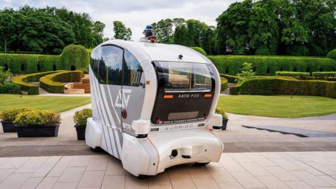 A driverless shuttle standing at the foreground in the middle of the shot. With green garden on the background. 