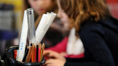 A generic photo shows anonymous primary school-age children in a classroom. In the foreground is a container full of pencils and rulers.
