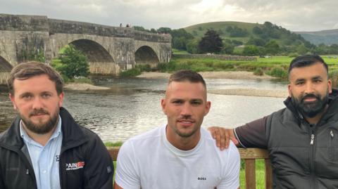 Nathan Clarkson, Perry Bruines and Safraz Hussain on a bench with the River Wharfe in the background. Mr Clarkson has short dark hair and short facial hair. He is wearing a black jacket. Mr Bruines has short, dark hair and is wearing a white t-shirt. Mr Hussain has short black hair with a black beard. He is wearing a black polo shirt and black gilet.
