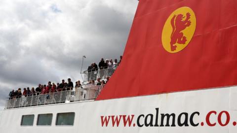 People standing on the upper deck of a CalMac ferry, with a grey sky in the background