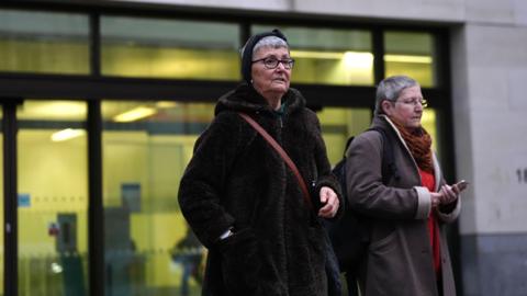 Diane Bligh, left, and Alyson Lee, right, standing outside court. Diane Bligh is wearing a furry jacket and a cross-body bag, while Alyson Lee wears a grey coat with a red jumper and russet scarf underneath. Both women have short grey hair and are wearing glasses.