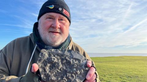 A man wearing a black beanie hat with pin badges smiles into the camera. He is holding a dark, textured rock containing lots of circular fossils. He is standing on a grassed area under a bright, blue sky. The sea is visible in the background.