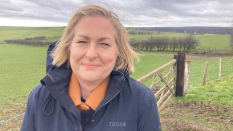 Alison Hume smiles at the camera, with some green fields behind her and blackened moorland in the distance.