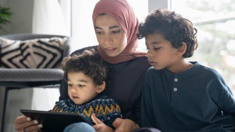 Stock image shows a mother holding a tablet while watching something on the screen with two young children at home in a living room.
