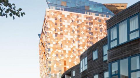 The Cube 25-storey building, taken from street level on the Birmingham Canal. The building is clad in brown and white tiles arranged in geometric patterns, and lower-rise flats can be seen on the right, clad in brown wood.