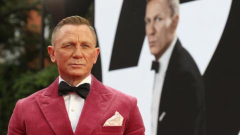Actor Daniel Craig in red tux stands in front of James Bond promotional poster