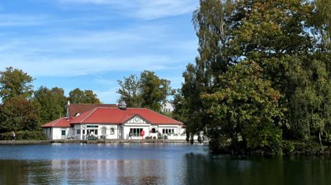 A photo of the boathouse at Rouken Glen Park. A white building with a red roof near a pond.