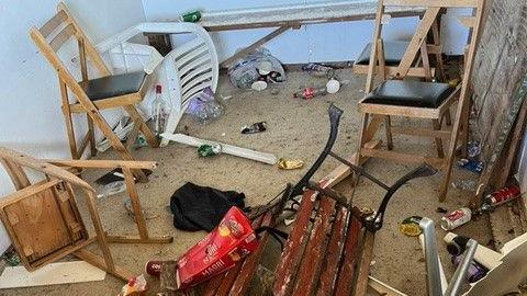 A disordered room with debris, featuring two wooden chairs with black seats, a broken bench with red slats and a black metal frame, an overturned white plastic chair, and an office chair. The floor is littered with rubbish including bottles, cans, food wrappers, and a broken piece of plasterboard.
