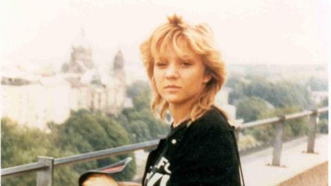 An old photograph of a young woman, standing on a balcony and looking at the camera. She has blonde hair and is wearing a black top. 