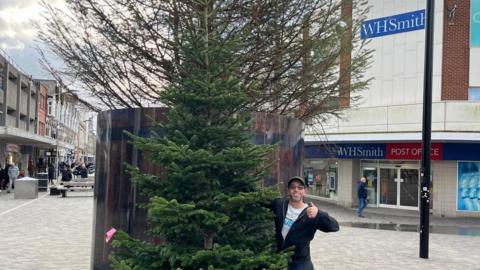 A ma is stood holding a green 13ft christmas tree. Behind him is the controversial High Street tree which has bare branches.