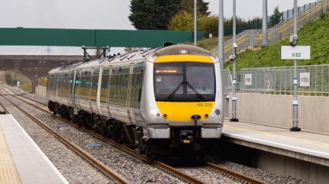 A yellow and silver multiple unit train stationary at the platform at Winslow station. There is a green-sided bridge above the platforms behind the train. A grass bank rises to the right alongside the platform.
