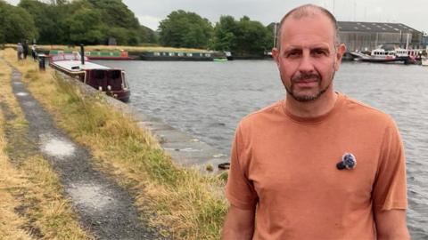 Mark Sadler stands in the foreground beside a wide waterway on an overcast day. He is wearing a light brown T-shirt and faces the camera. To the left, a narrow towpath runs alongside the water, bordered by dry yellow grass. Several narrowboats are moored along the bank, with more boats visible across the water. Trees line the far shore, and a large industrial-style building stands in the background.