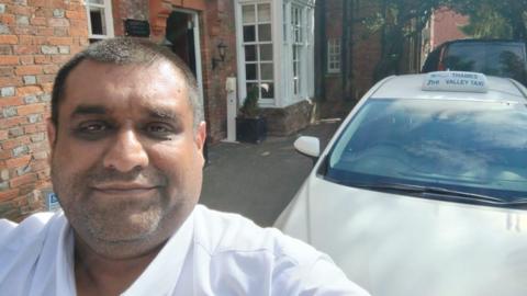 Yaseen Yusuf smiles for a selfie outside his home. He has a shaved head and light beard. He is wearing a smart white shirt and his white cab can be seen in the background. 